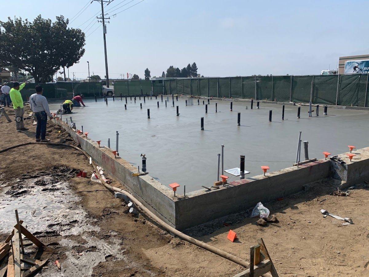A large poured foundation with column positions set into it, formed and screeded, with a construction fence behind.