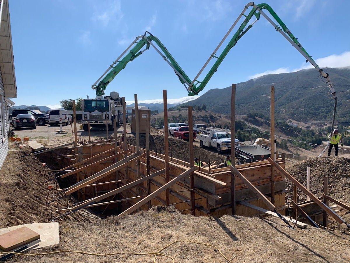 A green boom pump reaching over a hillside cut to place concrete into deep braced formwork, with the coastal hills behind.