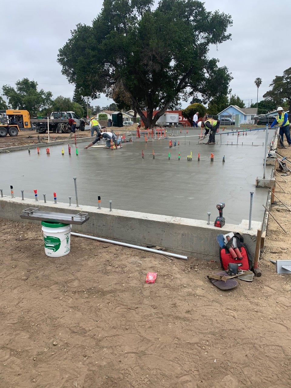 A large floor slab being floated flat by a crew, with the stem wall already poured along the near edge and markers set across the pour.